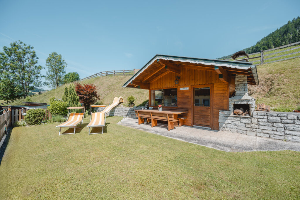 Deutsch: Ein gemütliches Gartenhaus umgeben von grünem Rasen und Büschen. Zwei Liegestühle laden zum Entspannen im Garten ein.
English: A cozy garden house surrounded by green grass and bushes. Two loungers invite you to relax in the garden.